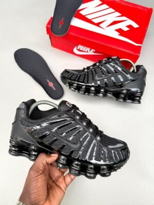 A pair of black and red Nike Air Max Plus sneakers with a red and white Nike shoe box on a wooden floor.
