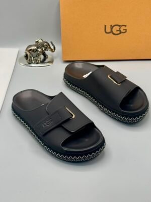 Grey UGG slide sandals with gold accents and stitched sole, displayed with a UGG box and decorative elephant figurine on a clean white surface.