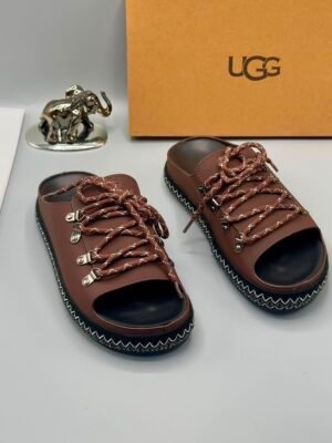 A pair of black leather UGG sandals with zigzag stitch and metal buckles, displayed with a UGG box and decorative elephant figurine.