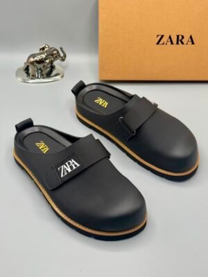 A pair of black leather clogs with yellow accents and ZARA logo, displayed with a brown shoe box and a small silver elephant statue on a white background.
